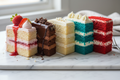Five cake slices in strawberry, chocolate, vanilla, blue velvet, and red velvet flavors displayed on a white marble surface